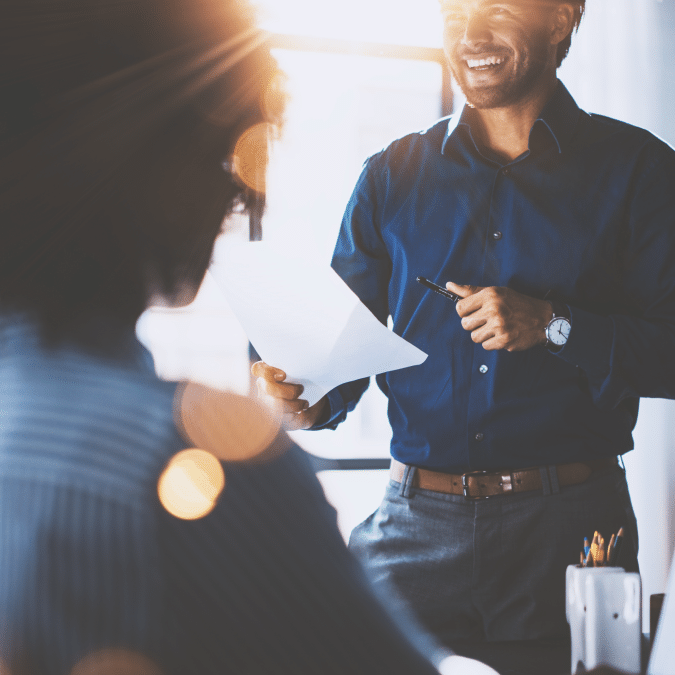 A man in a blue shirt stands holding papers and a pen, smiling as he talks to a seated person in an office setting with sunlight streaming in.