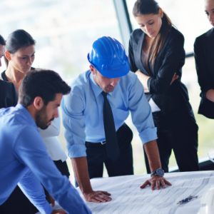 A group of professionals, including a person wearing a blue hard hat, closely examining blueprints on a table in a modern office setting.