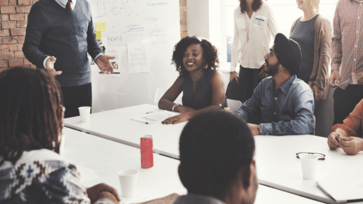 A diverse group of colleagues smiles and laughs during a meeting in a bright office. A man stands by a whiteboard with notes and diagrams, while others sit or stand around a table with drinks and papers.