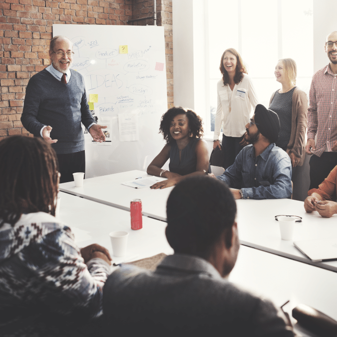 A diverse group of colleagues smiles and laughs during a meeting in a bright office. A man stands by a whiteboard with notes and diagrams, while others sit or stand around a table with drinks and papers.