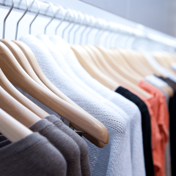 A row of wooden hangers holding various sweaters and shirts in neutral and warm colors on a clothing rack in a store or closet.