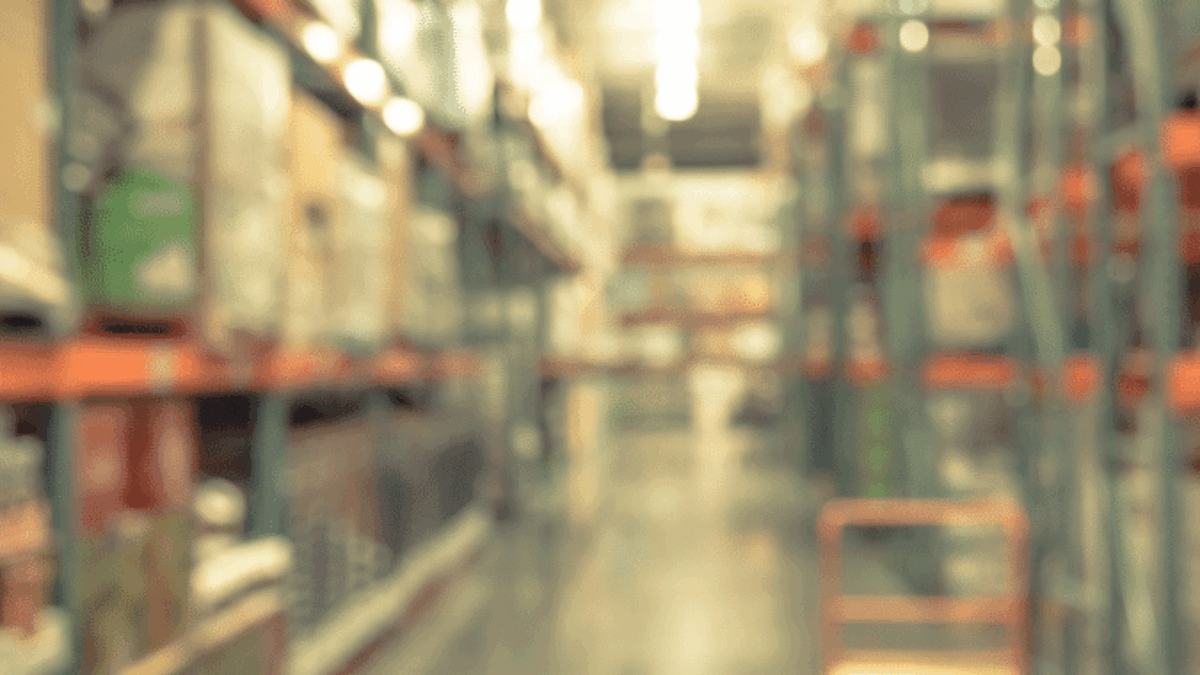 A blurred image of a warehouse aisle with shelves stacked with boxes and goods on both sides. A cart sits on the right side of the aisle under bright overhead lighting.