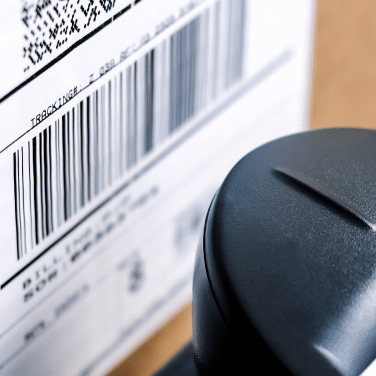 Close-up of a barcode and tracking label on a package, with part of a black handheld barcode scanner in the foreground.