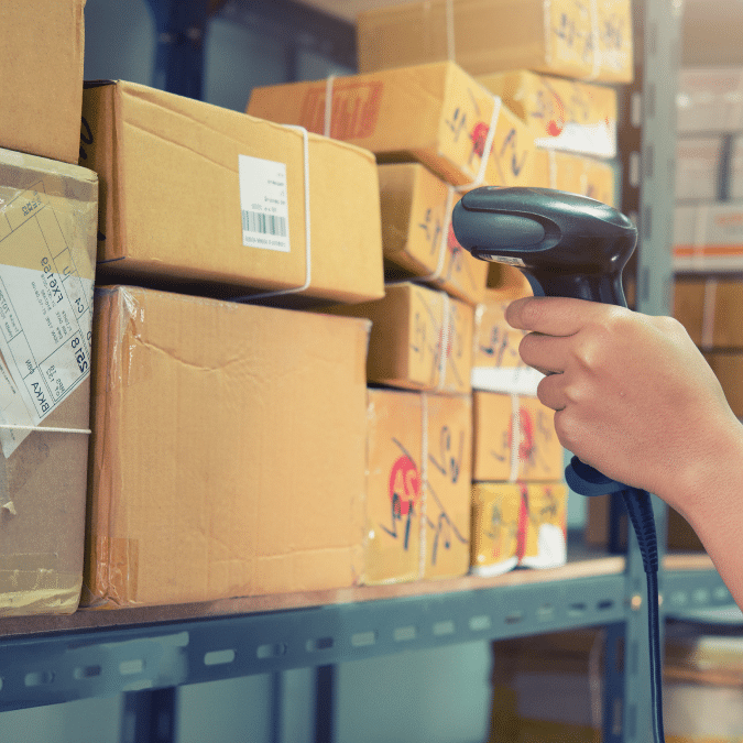 A person uses a barcode scanner to scan packages on metal shelves in a warehouse or storage area, surrounded by stacked cardboard boxes with shipping labels.