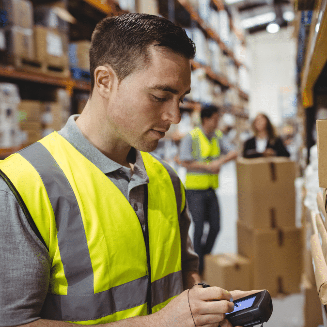 A warehouse worker in a yellow high-visibility vest scans a box with a handheld device, with shelves of packages and other workers visible in the background.