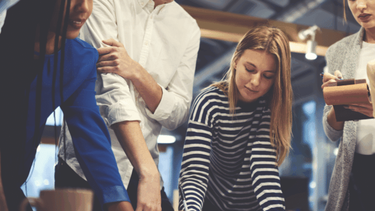 A group of young adults gathers around a table, collaborating and discussing documents in a modern office setting. They appear focused and engaged, with papers, notebooks, and a coffee cup visible on the table.