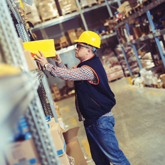 A warehouse worker in a yellow hard hat and safety vest stands by metal shelves, reaching to organize or retrieve a yellow box. The warehouse is filled with boxes and goods on racks.