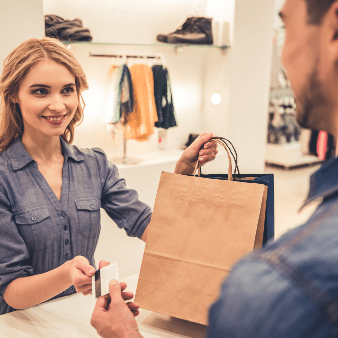 A woman at a retail counter smiles while handing a shopping bag to a customer, who is holding a credit card. Clothing and shoes are displayed on shelves in the background.