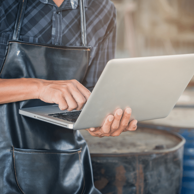 A person wearing a dark apron and plaid shirt uses a laptop, standing in what appears to be a workshop or industrial setting. Only their torso, arms, and the laptop are visible.