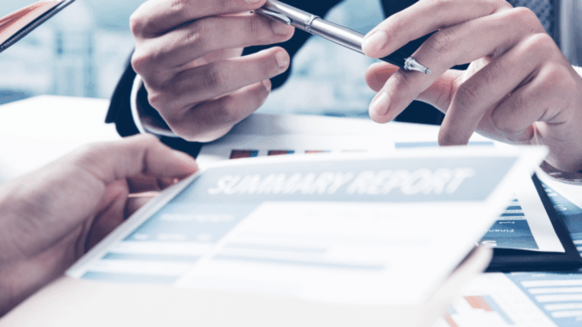 Two people in business attire discussing a summary report at a desk, with one person holding a pen and gesturing toward the document, suggesting a professional meeting or discussion.