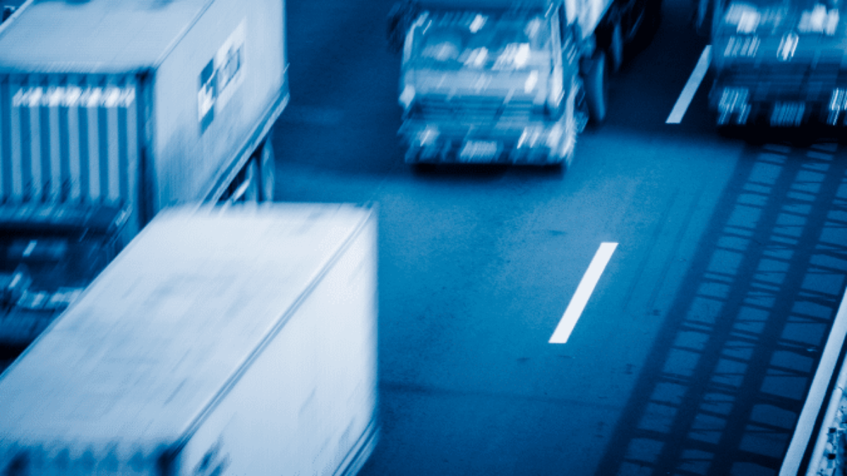 Several cargo trucks drive along a multi-lane highway. The image has a blue tint and appears to be taken with a motion blur effect, emphasizing the speed and movement of the vehicles.