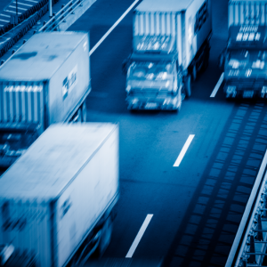 Several cargo trucks drive along a multi-lane highway. The image has a blue tint and appears to be taken with a motion blur effect, emphasizing the speed and movement of the vehicles.