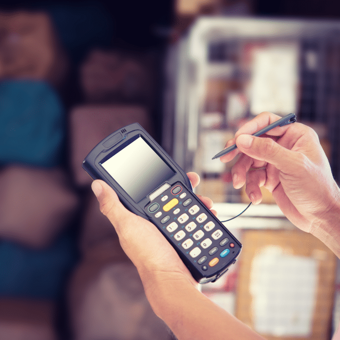 A person uses a stylus to operate a handheld electronic device with a keypad and screen, likely for inventory or data entry, in a warehouse or storage setting.