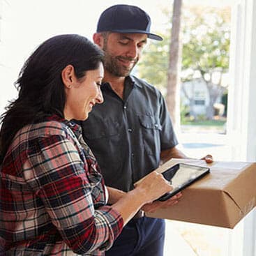 A woman signs for a package on a digital tablet handed to her by a delivery person wearing a cap and uniform, while standing at a door.