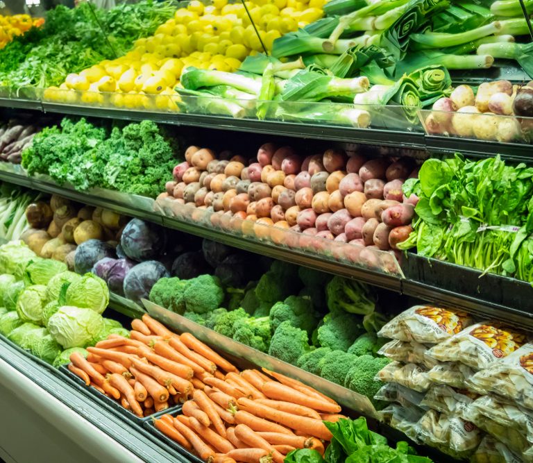 A grocery store produce section with neatly arranged vegetables including carrots, lettuce, broccoli, cabbage, potatoes, kale, squash, leeks, radishes, and packaged vegetables on multiple shelves.