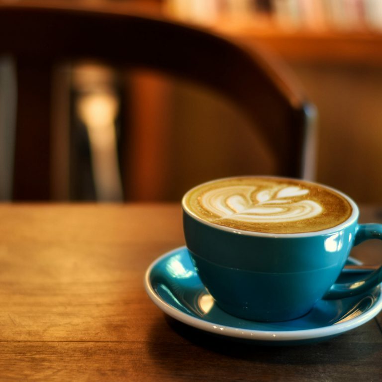 A blue cup and saucer with a latte featuring leaf-shaped latte art sits on a wooden table, capturing a cozy food and beverage moment against a blurred chair and warm-toned background.