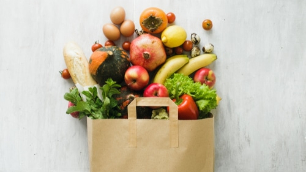 A paper bag filled with fresh groceries, including apples, bananas, pomegranate, eggs, tomatoes, lettuce, a loaf of bread, and a variety of vegetables, against a light background.