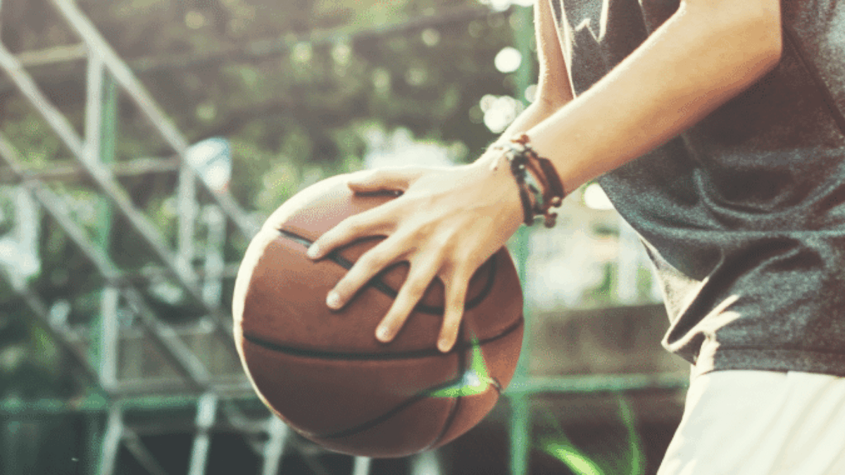 A person wearing a sleeveless shirt dribbles a basketball on an outdoor court, with sunlight filtering through trees in the background.