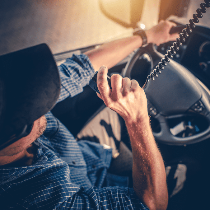 A person wearing a cap and a blue plaid shirt drives a vehicle while holding and speaking into a CB radio microphone, with sunlight shining through the window.