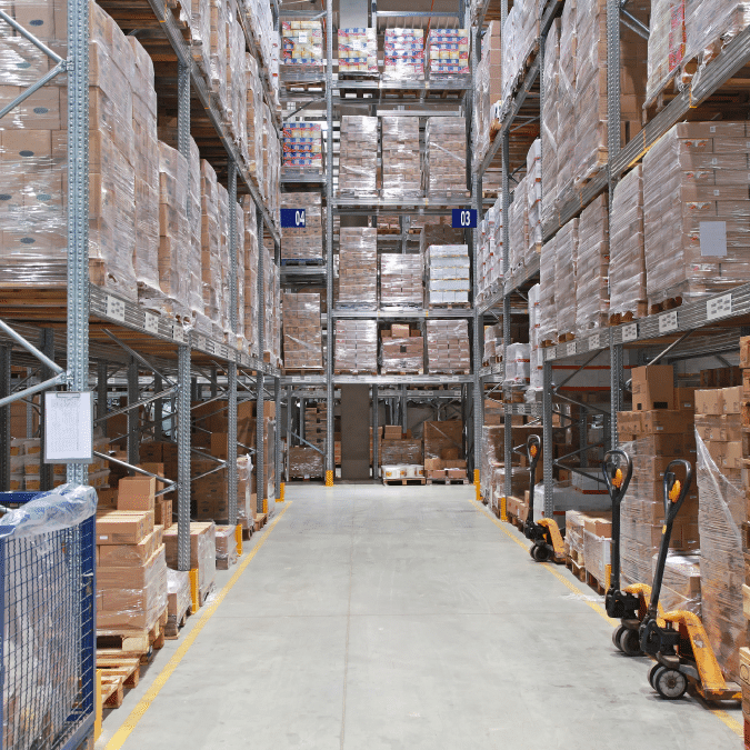A warehouse aisle with tall shelves stacked with pallets of boxed goods. Pallet jacks are visible on the right, and the floor is clear, allowing access for transport and movement.