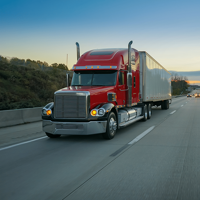 A red semi-truck with a large silver trailer drives on a highway at dusk, passing grassy roadside and trees under a clear blue sky.