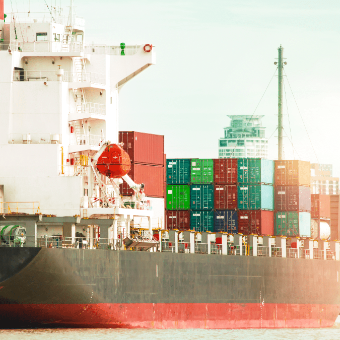 A large cargo ship loaded with colorful shipping containers docked at a port, with modern buildings visible in the background under a bright sky.