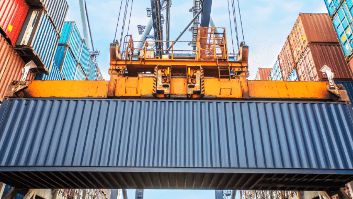 A large crane lifts a shipping container at a busy port, surrounded by stacked colorful containers under a blue sky.