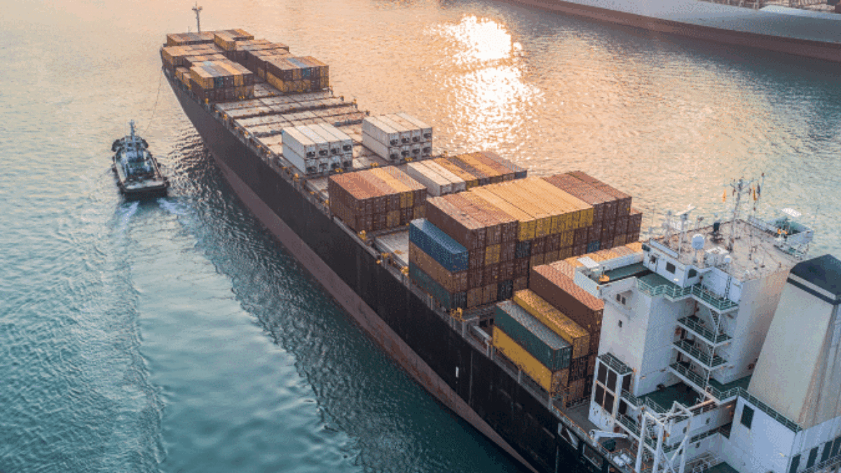 Aerial view of a large cargo ship loaded with colorful shipping containers sailing through a busy harbor, with cranes and container stacks visible along the port at sunset.