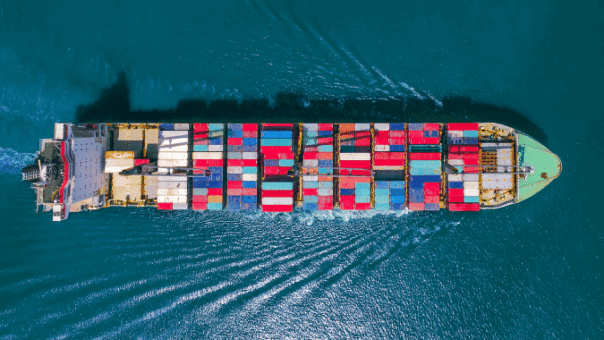 Aerial view of a large cargo ship carrying colorful shipping containers, sailing through calm blue ocean water, leaving a visible wake behind.