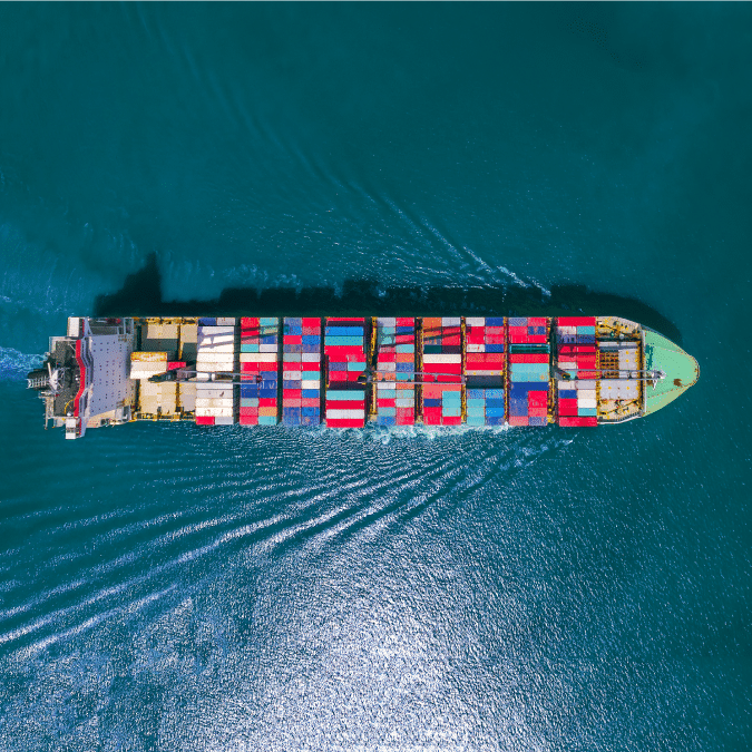 Aerial view of a large cargo ship carrying colorful shipping containers, sailing through calm blue ocean water, leaving a visible wake behind.