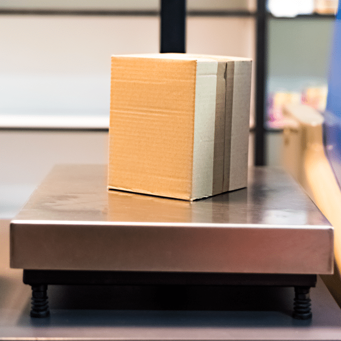 A small cardboard box sits on a metal platform or scale, possibly in a shipping or warehouse setting, with blurred shelves in the background.
