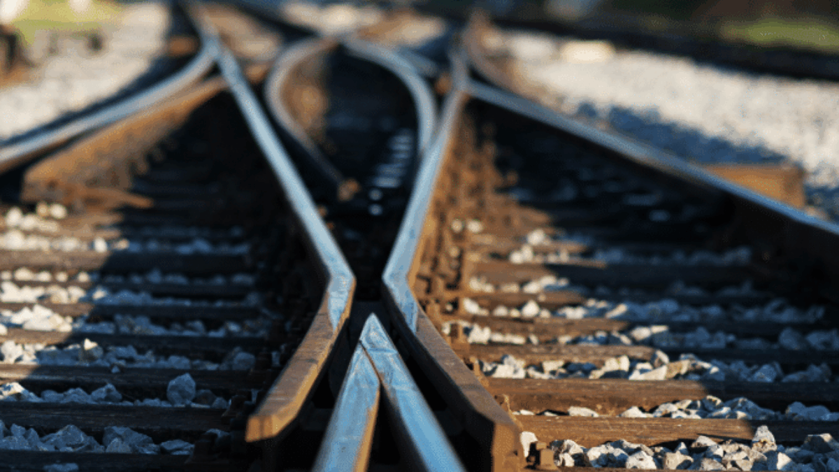 Close-up view of railroad tracks converging and splitting, with gravel between the rails and blurred landscape in the background on a sunny day.