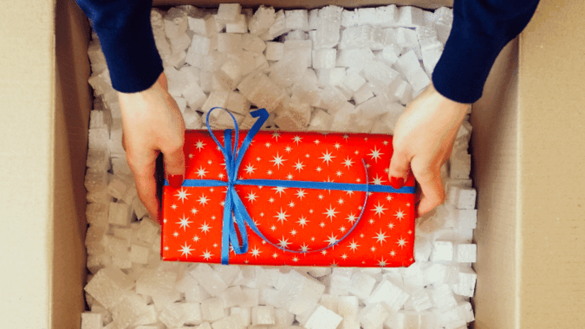 Person placing a red, star-patterned gift wrapped with a blue ribbon into a cardboard box filled with white packing peanuts, viewed from above.