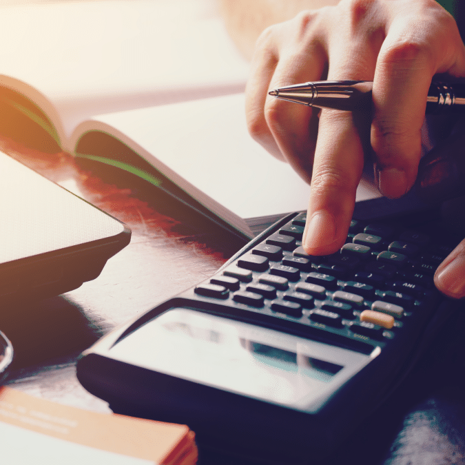 A person uses a calculator with one hand while holding a pen in the other, sitting at a desk with an open notebook, a laptop, and orange sticky notes nearby. Sunlight shines across the workspace.