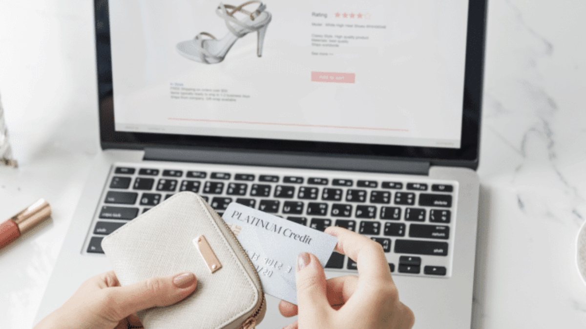 A person holds a wallet and a credit card while shopping for high-heeled shoes on a laptop, with an online shop website displayed on the screen.