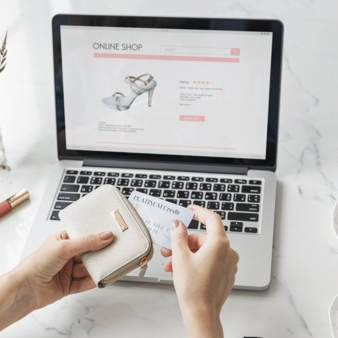 A person holds a wallet and a credit card while shopping for high-heeled shoes on a laptop, with an online shop website displayed on the screen.