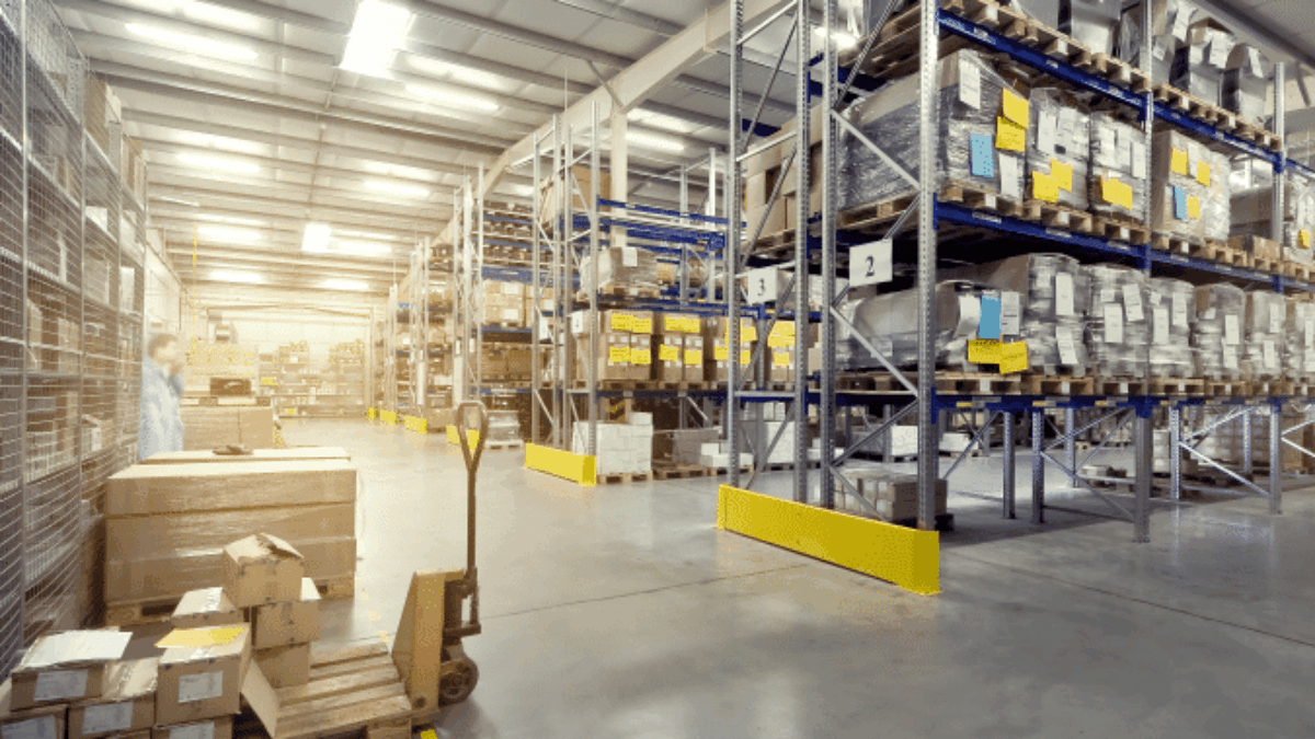 A spacious warehouse with high shelves filled with boxes and packages, a pallet jack loaded with boxes, and bright overhead lighting illuminating the clean, organized storage area.