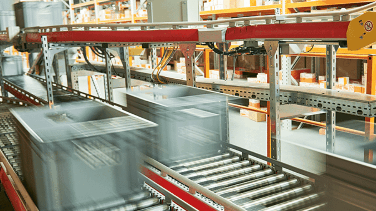 Automated conveyor system in a warehouse, designed with advanced systems engineering and automation, transports gray storage bins swiftly along metal rollers, while shelves stocked with orange and white boxes line the background.