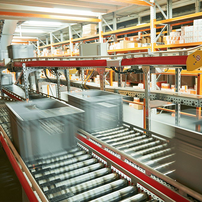 Automated conveyor system in a warehouse, designed with advanced systems engineering and automation, transports gray storage bins swiftly along metal rollers, while shelves stocked with orange and white boxes line the background.