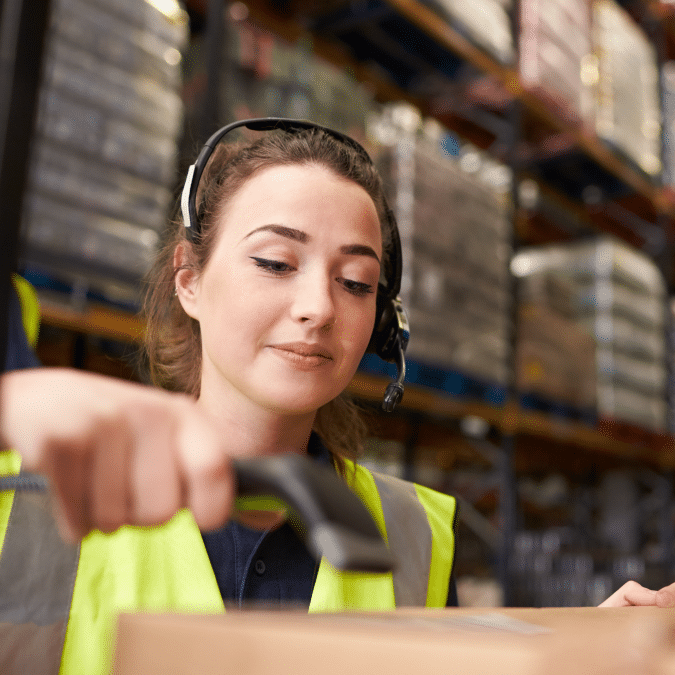 A woman wearing a headset and high-visibility vest scans a package with a handheld barcode scanner in a warehouse filled with shelves and boxes.