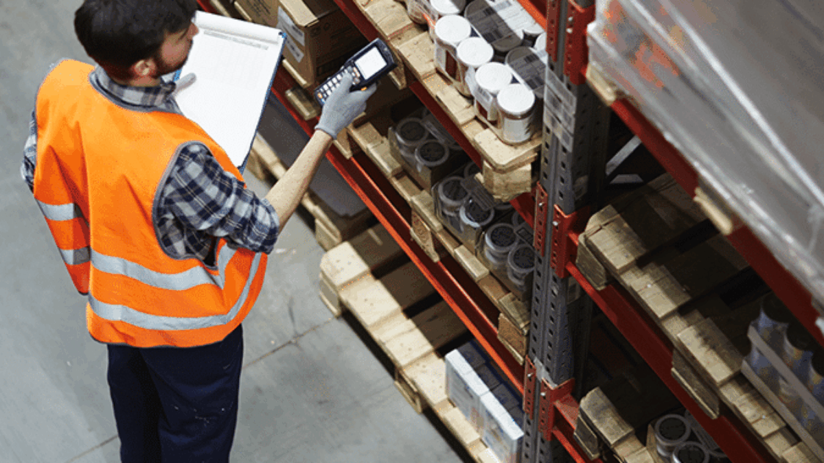 A warehouse worker wearing an orange safety vest and gloves uses a handheld scanner to check inventory on shelves, holding a clipboard and standing near stacks of boxed goods.