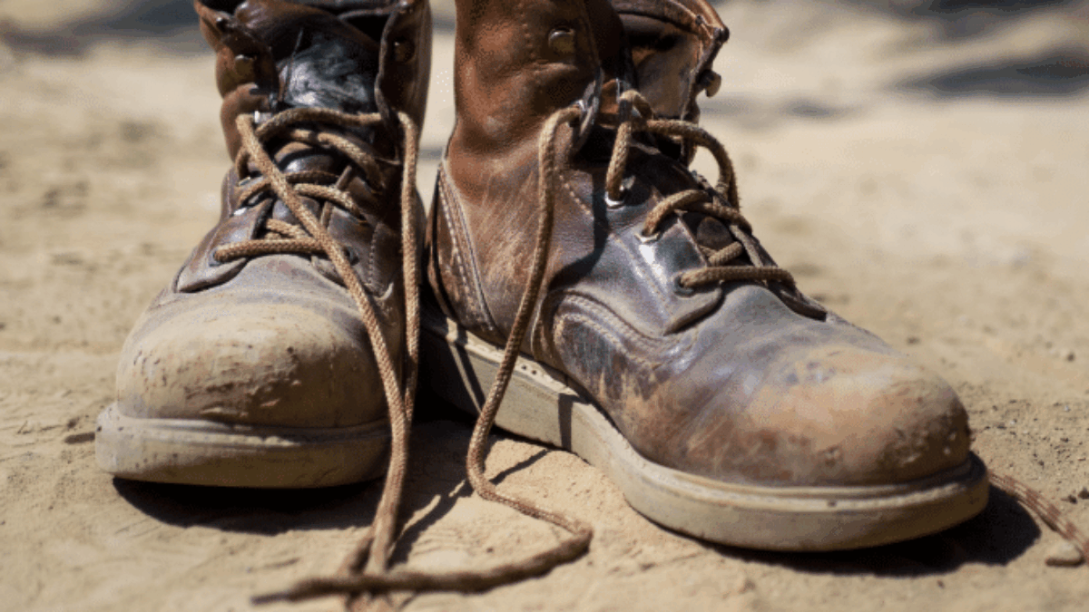 A pair of worn, brown leather work boots with untied laces sits on sandy ground outdoors, showing scuffs and signs of heavy use.
