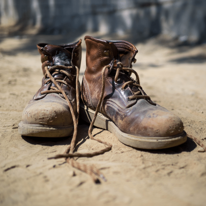 A pair of worn, brown leather work boots with untied laces sits on sandy ground outdoors, showing scuffs and signs of heavy use.