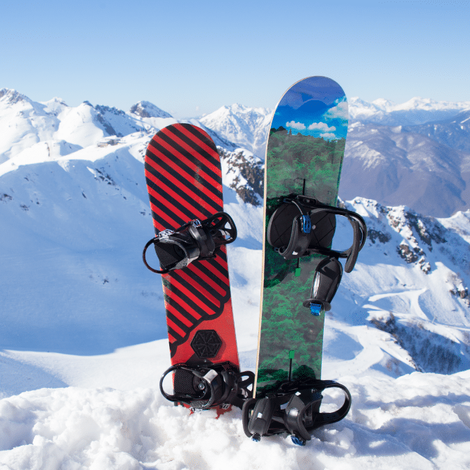 Two snowboards, one red with black stripes and one with a green and blue nature design, standing upright in the snow on a mountain with snowy peaks in the background under a clear blue sky.