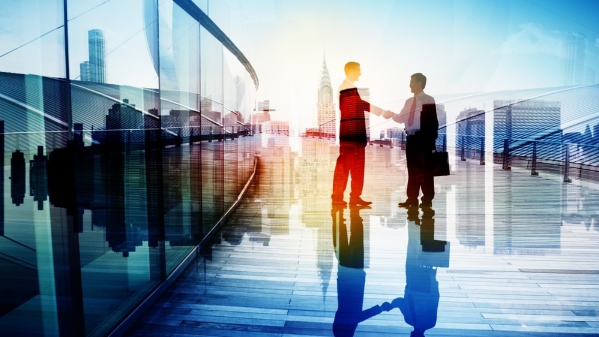 Two businesspeople shake hands on a modern glass walkway, with a city skyline and tall buildings visible in the background, and their reflections on the shiny floor.