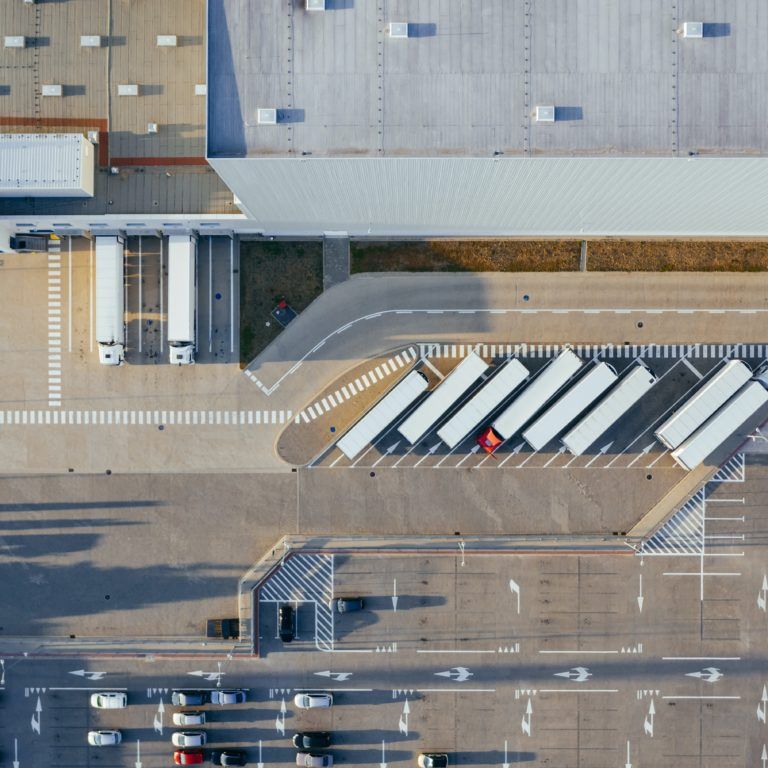 Aerial view of a warehouse with several trailers parked in loading bays, a few trucks at the docks, and a parking lot with cars in the foreground. The area is organized with marked lines and roads.