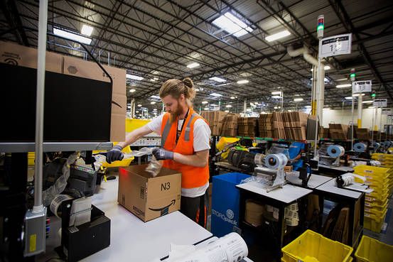 A worker wearing an orange safety vest packs a cardboard box at a workstation in a large warehouse filled with shelves, cardboard boxes, and packing materials.
