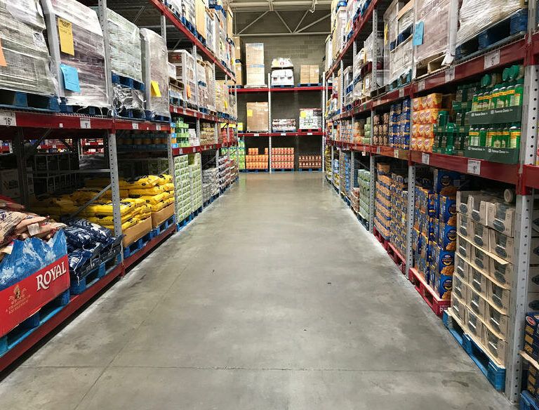 A wide aisle in a warehouse-style grocery store with shelves stacked high with bulk food items and large boxes on both sides. The concrete floor is clean and there are no shoppers visible.