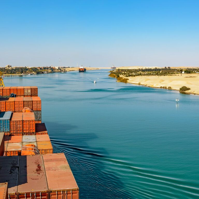 A cargo ship loaded with containers sails through the Suez Canal, bordered by sandy desert on one side and a distant cityscape on the other, under a clear blue sky.