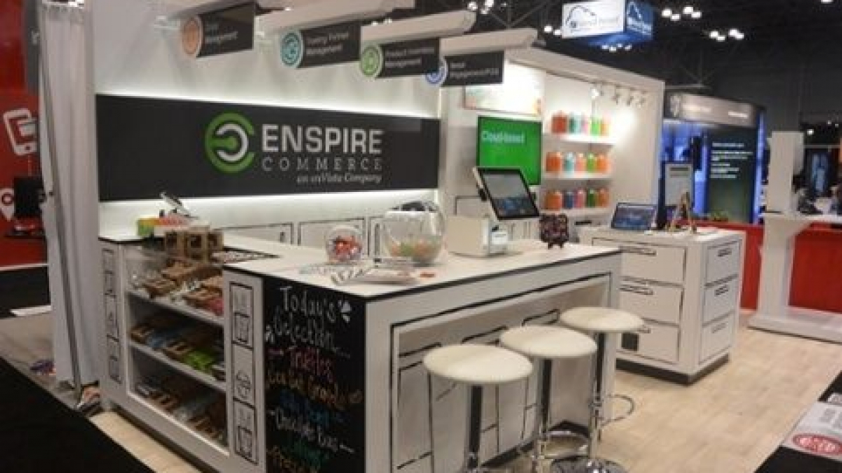 A modern trade show booth for Enspire Commerce features white counters, bar stools, computer screens, and colorful candy jars. Signage and product displays are arranged neatly throughout the space.